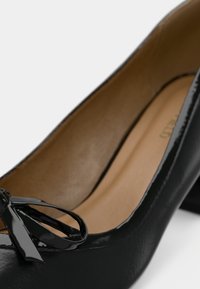 Μαύρη παλτό λουστρίνι γυναικεία παπούτσια με χαμηλό τακούνι και διακοσμητική φιόγκο στην άκρη, με μπεζ εσωτερική επένδυση.