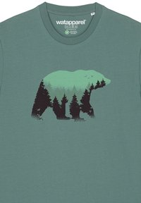 T-shirt en coton teal avec une silhouette d'ours noir remplie de graphiques de forêt verte et de ciel, représentant des arbres et des oiseaux.