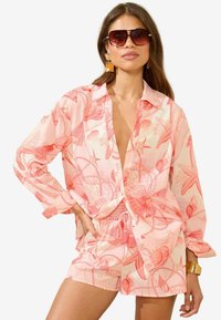 Roze overhemd met knopen en een patroon van schelpen en sterren, gecombineerd met bijpassende shorts met een trekkoord. Accessoires zijn onder andere oversized zonnebrillen.