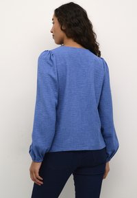 Blouse bleue légère avec des manches bouffantes et un tissu texturé. La vue de dos montre un col rond et une coupe ample.