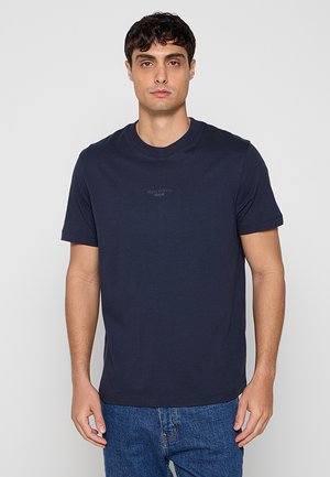 Mężczyzna z krótkimi ciemnymi włosami, ubrany w jednolity granatowy T-shirt i niebieskie dżinsy, stojący na jednolitym jasnoszarym tle.