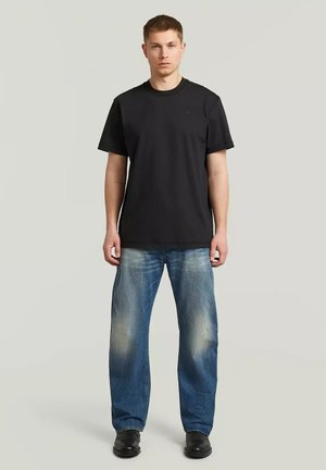 Jonge man die recht vooruit staat, draagt een effen zwart T-shirt, losse blauwe jeans met vervaagde plekken en zwarte schoenen tegen een neutrale achtergrond.