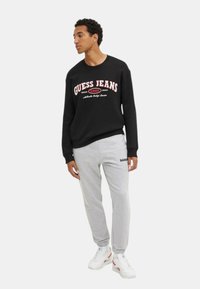 Sudadera negra con el logo de "GUESS JEANS" y pantalones de chándal grises. Presenta puños acanalados, corte relajado y detalles del logo en contraste en los pantalones.