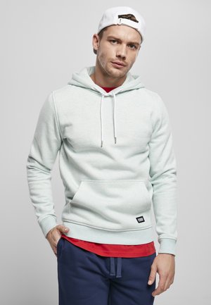 Haut à capuche vert menthe clair avec une poche kangourou, des cordons de serrage et des poignets côtelés. Superposé sur une chemise rouge, associé à un pantalon de jogging bleu marine.