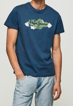 Man wearing a blue Pepe Jeans London T-shirt and light blue jeans, with left hand in pocket and right arm relaxed by side.