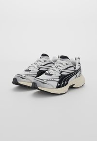 Chaussures de sport blanches et noires, en matériaux en mailles et synthétiques, à forme arrondie, avec des semelles texturées et des détails de design contrastés.
