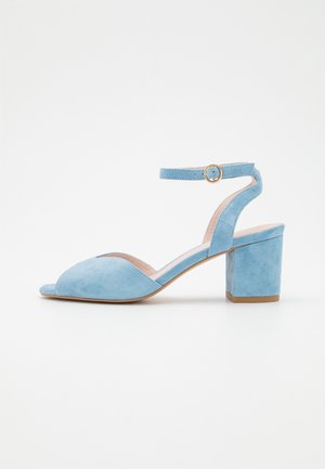 Hellblaue Veloursleder-Sandalette mit offenem Zehenbereich, Blockabsatz und Knöchelriemen mit einer kleinen goldenen Schnalle, dargestellt auf weißem Hintergrund.