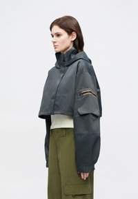 Siv skrajšan suknjič s kapuco, z žepom na rokavu z zadrgo in oranžnim poudarkom. Ima gladko, vodoodporno teksturo.