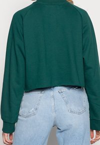 Felpa verde corta con lunghe maniche raglan, texture morbida e collo alto. Abbinata a jeans chiari strappati.