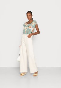 Top teal com estampado floral e mangas curtas, calças largas creme, sandálias de salto amarelas e uma pequena bolsa branca.