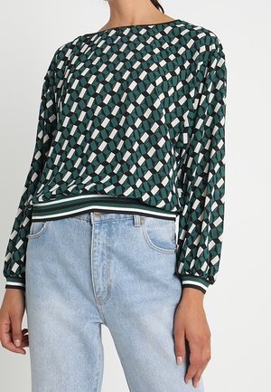 Langærmet grøn mønstret top med geometrisk design, stribede manchetter og kant, båret med lyseblå højttaljede jeans mod hvid baggrund.