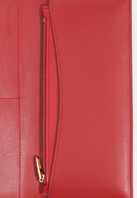 Czerwony skórzany kopertówka o fakturowanym wykończeniu, z złotym zamkiem błyskawicznym i eleganckim, zaokrąglonym otwarciem. Nazwa marki wytłoczona z boku.