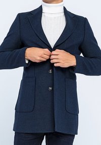 Marinblå ullrock med smal passform, med en knäpp krage, två framfickor och tre framknappar. Långa ärmar syns.