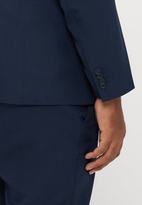 Marineblauwe colbertmouw met vier kleine knopen, gladde textuur, getailleerde pasvorm en een subtiele glans, gecombineerd met donkere broek.