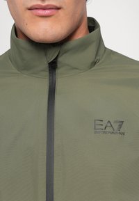Oliven grønn zip-jakke med høy krage, med svart glidelås og EA7-logo på venstre bryst. Glatt tekstur, lett materiale.