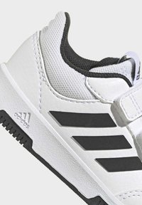 Biały but sportowy z fakturowaną cholewką ze skóry, czarną siateczkową podszewką i trzema czarnymi paskami po bokach. Wyposażony w pasek na rzep.