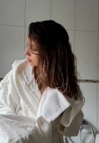 Zachte witte badjas met gevoerde textuur, voorzien van een sjawlboord en losse mouwen. Persoon met nat haar, gedeeltelijk zichtbaar, in een bathroom omgeving.