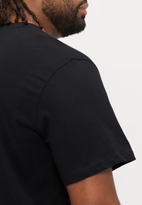 Μαύρο κοντομάνικο μπλουζάκι, κατασκευασμένο από μαλακό βαμβακερό ύφασμα, με λαιμόκοψη σε σχήμα κύκλου και άνετη εφαρμογή με ελάχιστες λεπτομέρειες ραφών.