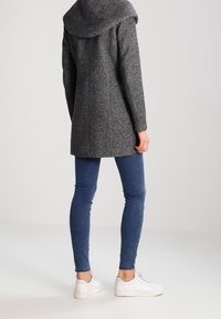 Abrigo de lana gris con una gran capucha, corte recto y tela texturizada. Se lleva sobre unos vaqueros azules y zapatillas blancas, mostrando un conjunto informal.