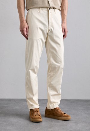 Beige bomuldsbukser med lige ben-design og to frontlommer, parret med brune ruskinds sneakers. Enkle, rene linjer og minimal detaljering.