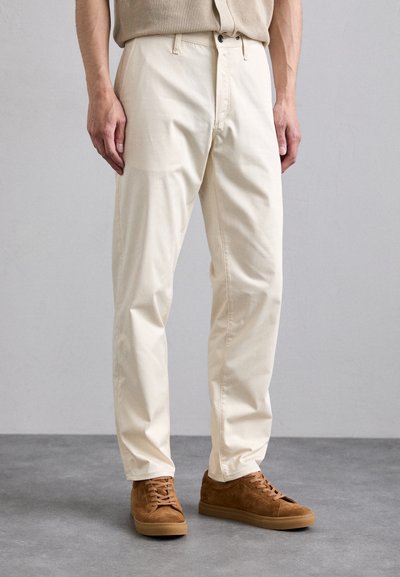 Pantalons en coton beige avec une coupe droite et deux poches avant, associés à des baskets en daim marron. Lignes simples et épurées, avec un minimum de détails.