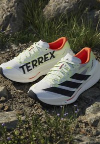 Ζευγάρι λευκά παπούτσια τρεξίματος Adidas Terrex με μαύρες ρίγες και πορτοκαλί εσωτερική επένδυση τοποθετημένα σε βραχώδες έδαφος με γρασίδι.