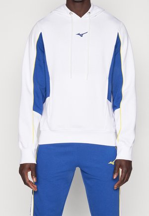 Mann trägt weißen Hoodie mit blauen und gelben Einsätzen und passende blaue Hose mit gelbem Rand und kleinen Logos auf Brust und Oberschenkel.
