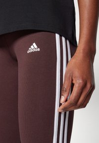 Adidas leggings i mörk burgund med vita sidostreck. Närbild av tygtextur och logotyp placerad på låret, i kombination med en svart topp.