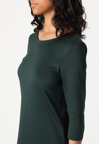 Nainen, jolla on tummat kiharat hiukset, pukeutuneena tummanvihreään pitkähiharaiseen paitaan, kuvattuna hartioista alas keskivartaloon, kasvot vasemmalle.