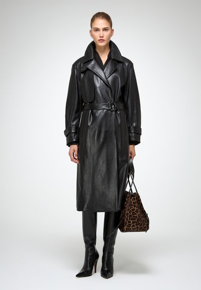 Donna che indossa un trench coat in pelle nero con cintura, stivali neri con tacco alto, mentre tiene una borsa con stampa leopardata, in piedi su uno sfondo neutro.