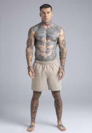 Mišičast moški z velikimi tetovažami na prsih, rokah in nogah, oblečen v bež kratke hlače in srebrne ogrlice, stoji bos na sivem ozadju.