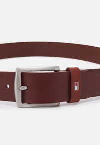 Ceinture en cuir marron avec une boucle en métal argenté, comportant des trous pour l'ajustabilité et un petit accent de marque avec des détails rouge et blanc.