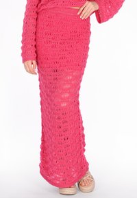 Frau trägt einen langen rosa gehäkelten Rock mit passendem Oberteil und beigen Plateausandalen, die Hand auf der Hüfte abgestützt.