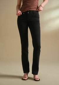Jeans de mezclilla negros con corte recto, diseño de cinco bolsillos, botón de metal y cremallera, combinados con una blusa entallada marrón y sandalias negras.