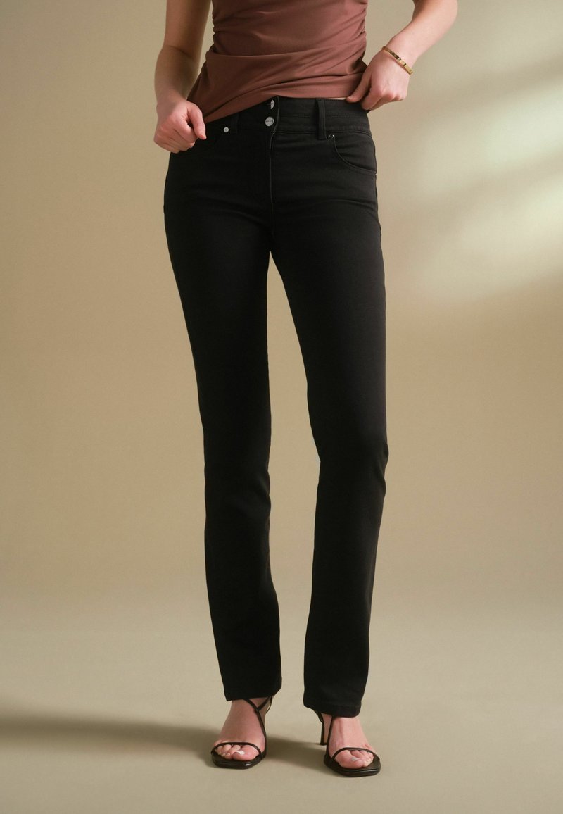 Jeans de mezclilla negros con corte recto, diseño de cinco bolsillos, botón de metal y cremallera, combinados con una blusa entallada marrón y sandalias negras.