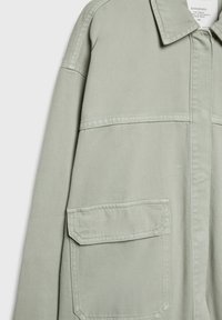 Jasnoszara kurtka dżinsowa z kołnierzem, widoczną kieszenią na klapie oraz szwami na przedzie rękawów i w okolicy klatki piersiowej.