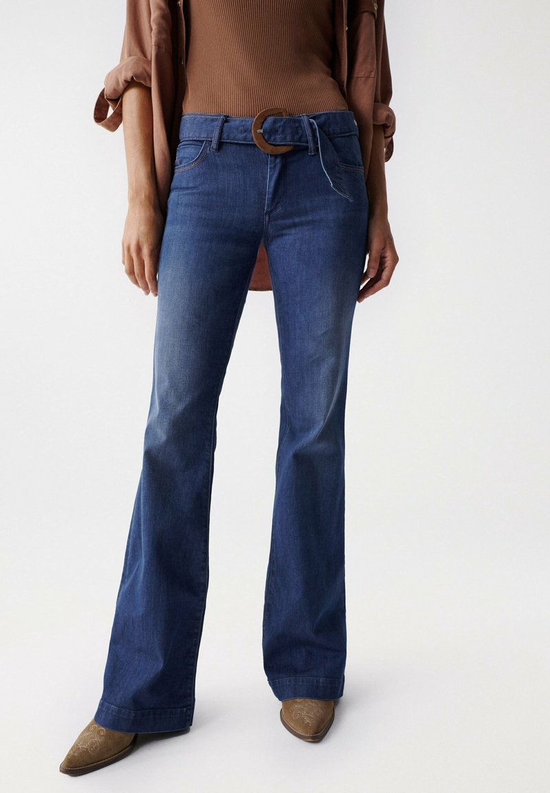 Mörkblå utsvängda jeans i denim, med ett brunt bälte med ett runt spänne. Slät textur och raka ben.