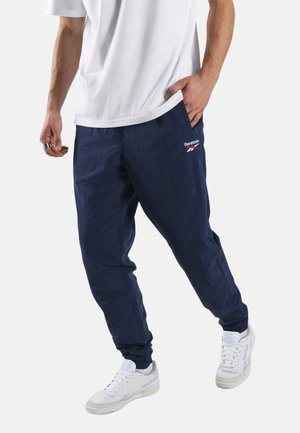Man draagt witte sneakers, marineblauwe Reebok trainingsbroek en een wit t-shirt, met één hand in de zak, tegen een effen achtergrond.