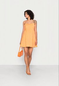 Mulher com cabelo encaracolado, vestindo um vestido laranja sem mangas, segurando uma bolsa laranja e usando sandálias de salto laranja, de pé contra um fundo branco.