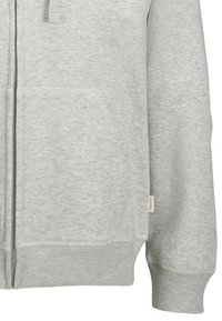 Haut à capuche zippé gris en tissu doux, avec une poche avant, des poignets et un ourlet côtelés, et une petite étiquette de marque sur le côté.