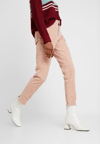 Pantalon en velours côtelé rose à coupe slim, avec poches latérales et fermeture éclair à l'avant. Porté avec des bottines blanches et un pull bordeaux.