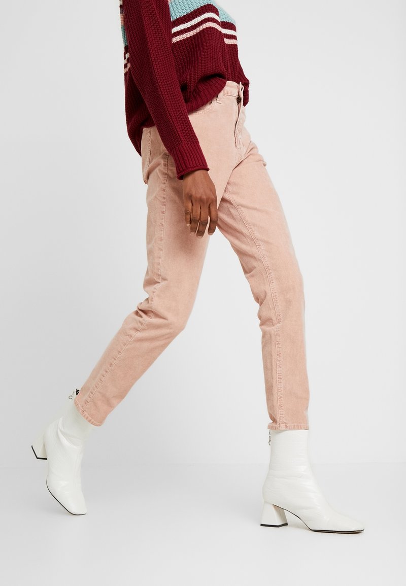 Pantalon en velours côtelé rose à coupe slim, avec poches latérales et fermeture éclair à l'avant. Porté avec des bottines blanches et un pull bordeaux.