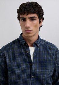 Camisa de botones de cuadros en azul marino y verde, hecha de tela suave. Cuenta con un collar puntiagudo, un bolsillo en el pecho y botones color marrón claro.