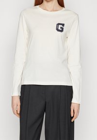 Langärmliges cremefarbenes T-Shirt aus weicher Baumwolle, mit einem marineblauen Logopatch auf der linken Brust und einem Rundhalsausschnitt. Kombiniert mit dunklen Hosen.