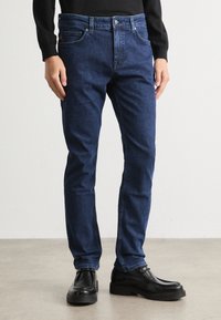 Temno modri kavbojke z ravno nogo, diskretno šivanje in gumbom za zapiranje, v kombinaciji s črnimi usnjenimi čevlji.