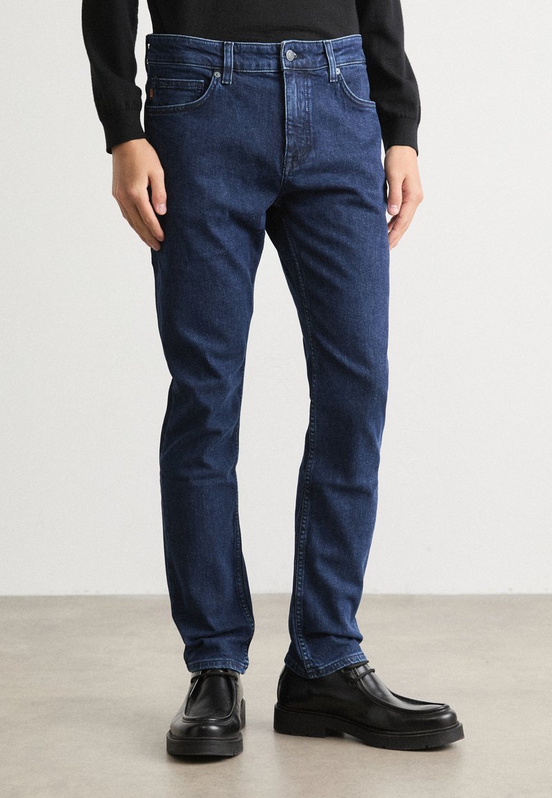 Temno modri kavbojke z ravno nogo, diskretno šivanje in gumbom za zapiranje, v kombinaciji s črnimi usnjenimi čevlji.