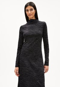 Femme aux longs cheveux noirs portant une robe longue à manches longues moulante à rayures zébrées noir et gris avec un col haut sur fond blanc.