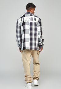 Camicia in flanella a quadri bianco e nero con una grande stampa "SILK" sul retro, abbinata a pantaloni cargo beige chiaro e sneakers bianche.