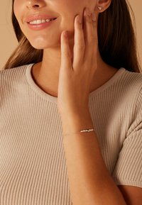 Bracelet en argent avec des perles blanches et noires, porté au poignet. Modèle avec des cheveux châtain clair, portant un t-shirt en maille beige, souriant doucement.