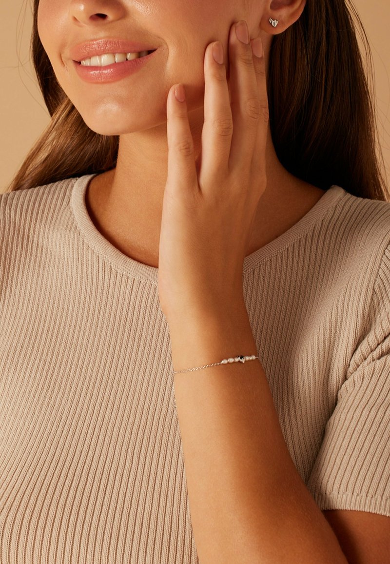 Bracelet en argent avec des perles blanches et noires, porté au poignet. Modèle avec des cheveux châtain clair, portant un t-shirt en maille beige, souriant doucement.